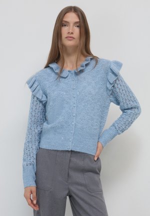Jeune femme portant un cardigan tricoté à volants bleu clair avec des motifs complexes et un pantalon gris taille haute, posant avec une main sur la hanche.
