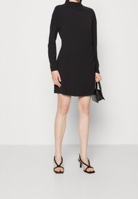 Femme portant une mini-robe noire à manches longues, des talons noirs à brides, tenant un petit sac à main noir devant un fond clair uni.