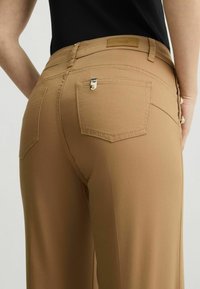 Bruine, stretchy broeken met een middelhoge tailleband, twee achterzakken en metallic hardware. Glad textuur met een strak ontwerp.