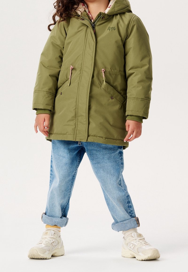 Parka vert olive avec capuche, deux poches avant, détails à cordon de serrage, et un intérieur doublé de tissu fleuri. Associée à un jean bleu retroussé et des baskets blanches.