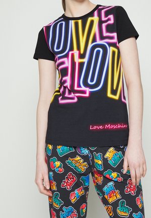 Person iført sort T-shirt med korte ærmer med neonlysende "LOVE"-tekst og sorte bukser dækket af farverige graffiti-stil logoer og sætninger.