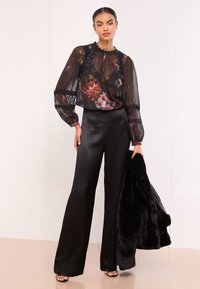 Žena oblečená v priesvitnej kvetinovej blúzke s čipkovými detailmi, čierne saténové nohavice so širokými nohami, čierne sandále, drží čierny kabát z umelú kožušinu.