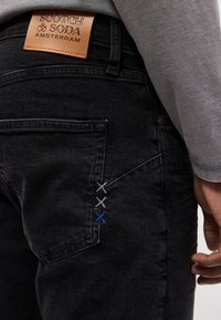 Πίσω τσέπη μαύρου τζιν με τρεις μικρές χρωματιστές ραφές και ταμπέλα από δέρμα ταμπά με την επιγραφή "Scotch & Soda Amsterdam."