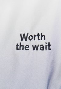 Tessuto bianco con testo ricamato nero che recita "Vale la pena aspettare." Texture liscia con una leggera lucentezza. Design semplice e minimalista.