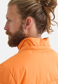 Chaqueta acolchada naranja con un cuello alto y textura acolchada. Presenta bolsillos con cremallera y un exterior suave. Modelo mostrado de perfil.