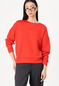 Felpa rossa over-size con scollo rotondo e spalle ribassate. Tessuto morbido e maniche lunghe e aderenti. Abbinata a pantaloni scuri.