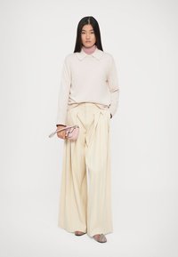 Maglione a maglia beige chiaro con colletto, pantaloni larghi crema con pieghe, tiene una piccola borsa rosa, indossa scarpe con motivi.