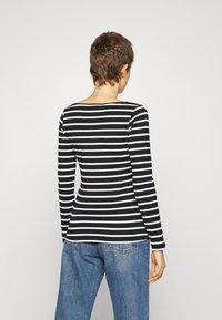 Person mit kurzen braunen Haaren, die ein schwarz-weiß gestreiftes Langarmshirt und eine blaue Jeans trägt, von hinten vor weißem Hintergrund.