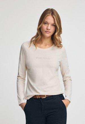Beige Langarmoberteil mit rundem Ausschnitt, versehen mit dem dezenten Text "Polo Club". Kombiniert mit dunklen Hosen und einem braunen Gürtel.