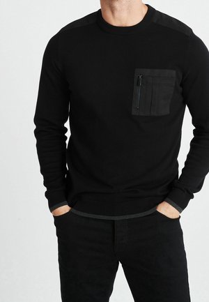 Homme portant un pull noir à manches longues avec une poche zippée sur la poitrine, les mains dans les poches, devant un fond clair uni.