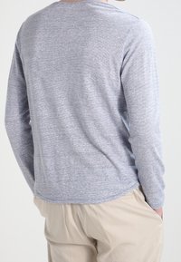 Chemise à manches longues gris clair avec une texture subtile, dotée d'un ourlet arrondi et d'un col rond. Portée avec un pantalon beige.