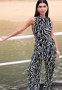 Zwart-wit jumpsuit met een zebraprint, mouwloos ontwerp, wijde pijpen en een gladde textuur. Model poseert bij water.
