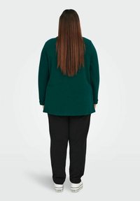 Žalias ilgomis rankovėmis marškinėlis su išlenktu kraštu, derinamas su juodais džinsais. Modelis stovi atgal, demonstruodamas laisvą prigludimą ir glotnią tekstūrą.