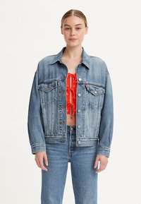 Giacca di jeans azzurro chiaro con vestibilità rilassata, chiusura a bottoni, tasche sul petto e cuciture a contrasto; indossata sopra un top rosso corto.