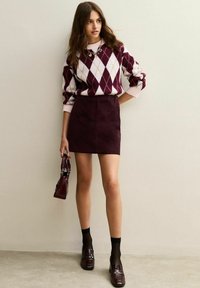 Bordeaux ruitjestrui met lichtroze accenten, gecombineerd met een bijpassende bordeaux corduroy rok. Zwarte sokken en glanzende loafers maken de look compleet.