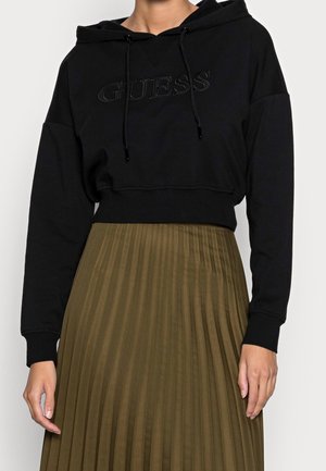 Persoon draagt een zwarte cropped hoodie met het "GUESS"-logo en een olijfgroene geplooide rok, staand tegen een effen lichte achtergrond.