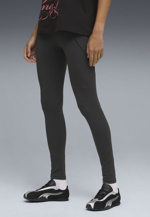 Persona que lleva leggings negros, zapatillas blancas y negras, calcetines blancos y una camiseta negra con texto rosa, de pie frente a un fondo gris.