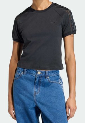 Mujer que lleva una camiseta negra corta con mangas de malla y vaqueros azules de tiro alto con cierre de botón y cremallera.