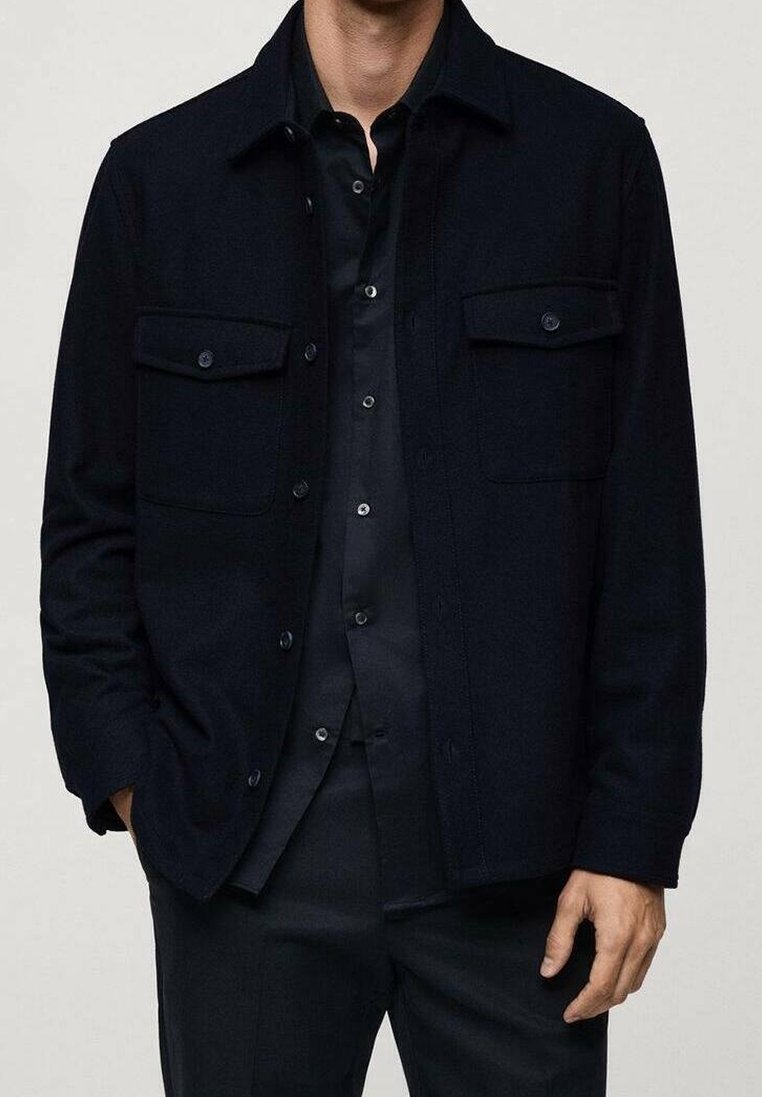 Homme portant une chemise noire boutonnée sous une veste noire avec des poches à rabat, debout avec une main dans la poche sur un fond uni.