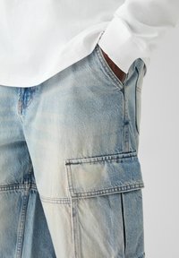 Světle modré cargo kalhoty vyrobené z denimu, s velkými bočními kapsami, viditelným prošíváním a volným střihem. Tkanina vykazuje mírné vyblednutí.