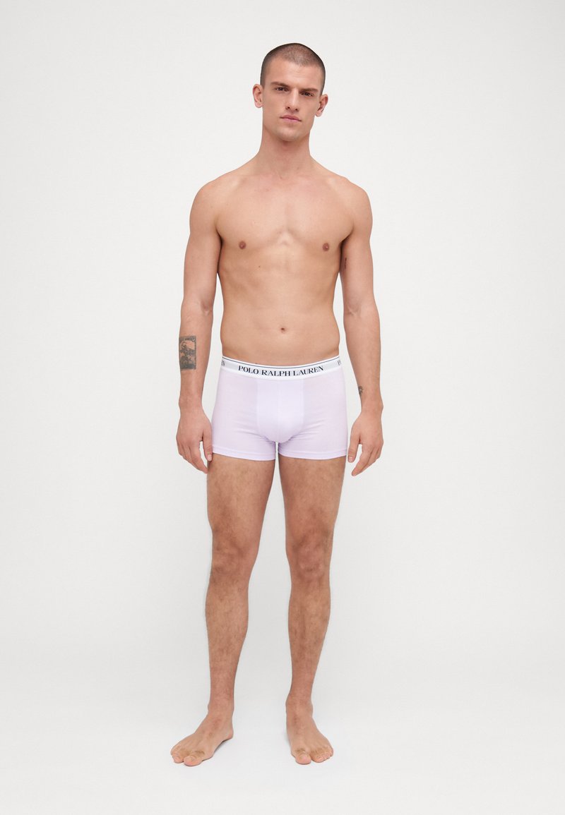 Boxer-briefs blancs avec une ceinture logo noire. Tissu lisse, design ajusté et sans motifs visibles. Le modèle se tient pieds nus devant un fond uni.