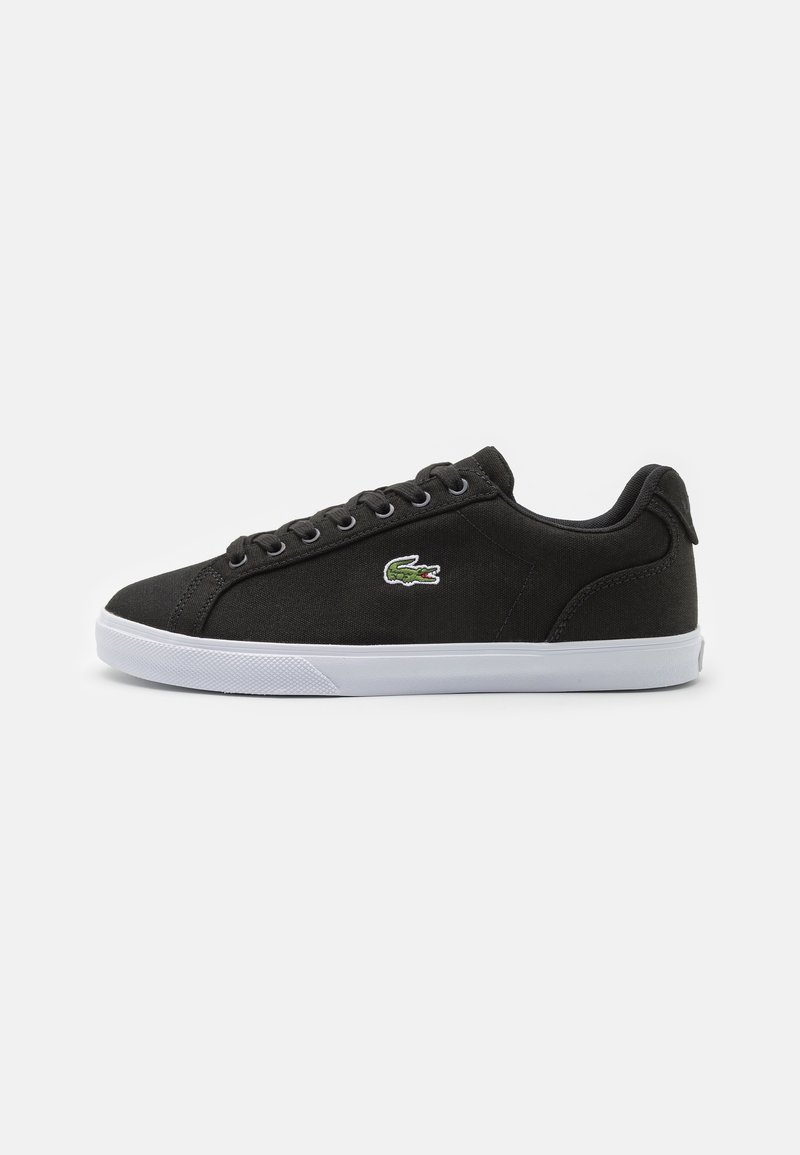 Sneakers bas noirs avec semelle blanche, lacets noirs et logo de crocodile vert sur le côté, sur un fond blanc uni.
