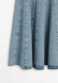 Lichtblauwe denimrok met een uitlopende pasvorm en verticale naaddetails, hangend tegen een witte achtergrond.