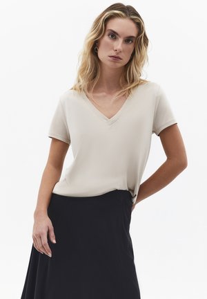 Vrouw met blond haar die een beige top met halve mouwen en V-hals draagt, ingestopt in een hooggetailleerde zwarte rok, staand met één hand op haar heup.