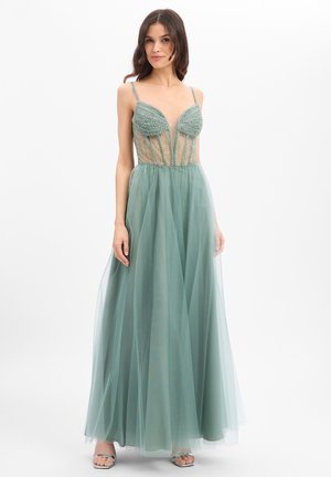 Vrouw draagt een vloerlange zeeschuimgroene jurk met kralenversierd lijfje en tüll rok, staand tegen een effen witte achtergrond.