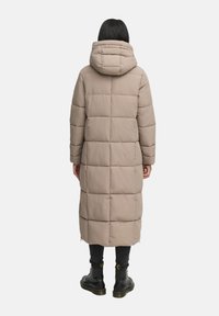 Abrigo de plumas largo color beige con capucha, que presenta un diseño acolchado y cremalleras laterales. Se lleva con pantalones oscuros y botas negras de cordones.