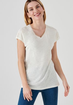 Korte mouw, crèmekleurig V-hals shirt met kantdetails op de schouders en een textuurstof. Gecombineerd met blauwe jeans, casual gedragen.