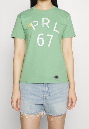 Personne portant un t-shirt vert avec le texte "PRL 67" et un short en jean bleu clair aux ourlets effilochés, debout devant un fond uni.