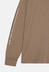 Brun langermet skjorte med elastiske mansjetter. Har hvit "NEW YORK CITY" tekst på ermet og et mykt, strukturert stoff.