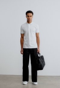 Λευκό πόλο πουκάμισο, μαύρο παντελόνι cargo, λευκά αθλητικά παπούτσια και μαύρη τσάντα. Το μοντέλο στέκεται μπροστά σε φωτεινό φόντο με ουδέτερη επένδυση δαπέδου.