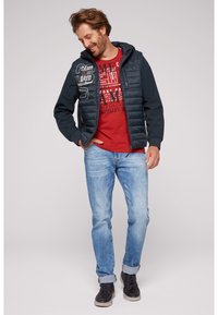 Jachetă albastră matlasată cu glugă închisă, tricou grafic roșu, blugi deschiși la culoare și adidași negri, având fermoar și detalii brodate.