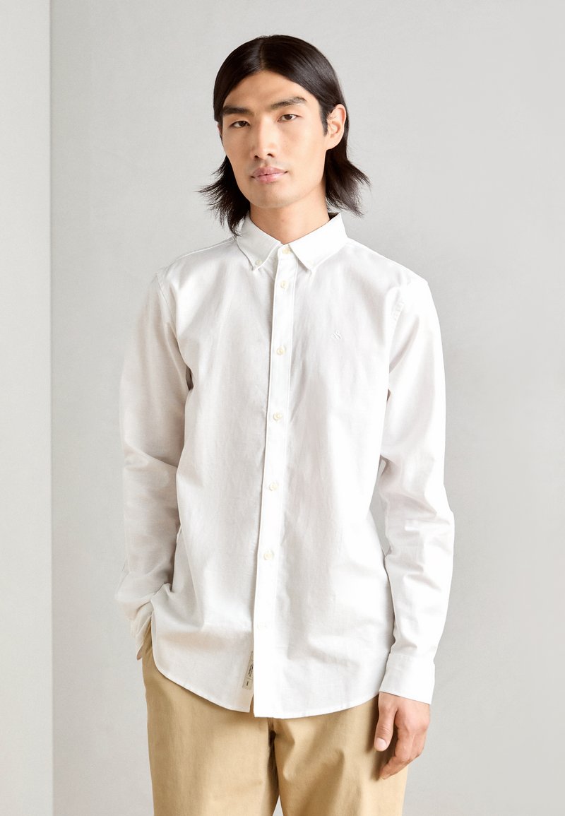 White button-up shirt made of cotton with a soft texture. Features a classic collar, long sleeves, and a subtle embroidered logo on the chest.