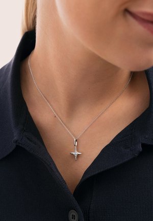 Collar de plata con un colgante en forma de estrella que tiene una gema central, colgando de una cadena delicada, usado sobre una camisa de punto oscura con cuello.