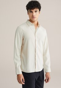 Svetlo béžová dlhá košeľa s golierom na zapínanie, jemná štruktúra a hladký povrch. Obsahuje jemné vertikálne pruhy a zapínanie na gombíky vpredu.