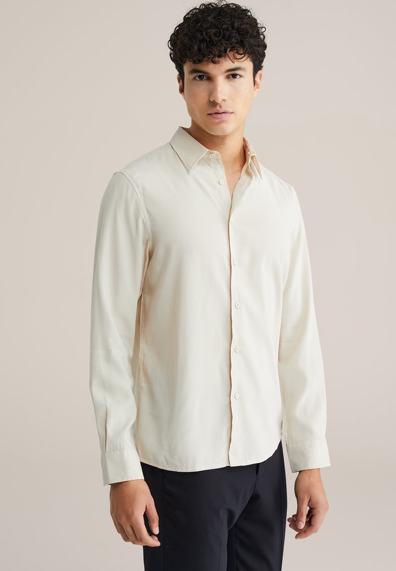Svetlo béžová dlhá košeľa s golierom na zapínanie, jemná štruktúra a hladký povrch. Obsahuje jemné vertikálne pruhy a zapínanie na gombíky vpredu.