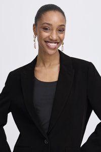 Ženska, ki se smehlja, oblečena v črni blazer, črno majico in viseče uhane s svetlimi kamni, na enobarvni svetli podlagi.