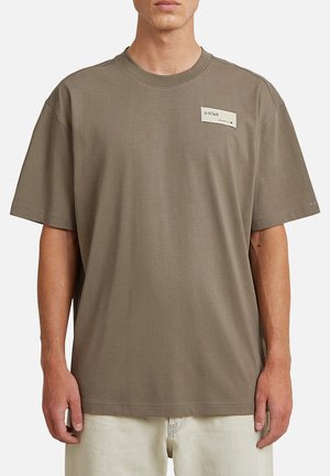 Mann trägt oversized olivgrünes G-Star T-Shirt mit kurzem Arm und Logopatch auf der Brust sowie helle Hose.