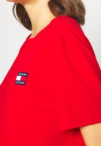 Osoba ubrana w jaskrawoczerwoną, oversize'ową koszulkę z małą naszywką z logo Tommy Jeans na piersi, widoczna od szyi do połowy torsu.