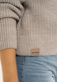 Grijze gebreide trui met een gestructureerd patroon, voorzien van een geribbelde zoom en een leren label met de tekst "Knit Factory" nabij de taille.
