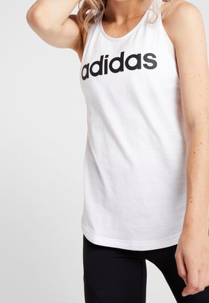 Žena v bílem tílku Adidas bez rukávů a černých legínách, zvednutá ruka k hlavě, na jednoduchém světlém pozadí.