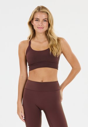 Frau mit langen blonden Haaren, die einen passenden dunkelbraunen Sport-BH und Leggings trägt, steht vor einem schlichten hellen Hintergrund.