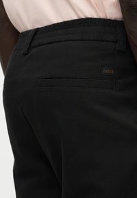 Κοντινό πλάνο μαύρου tailored παντελονιού με πίσω τσέπη και μικρή ορθογώνια ετικέτα της μάρκας BOSS κοντά στη μέση.