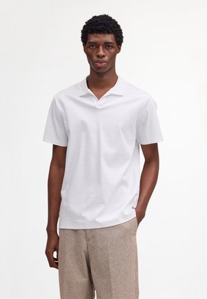 Jeune homme aux cheveux courts et bouclés portant un polo à manches courtes blanc et un pantalon beige texturé, sur fond blanc uni.