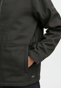 Veste noire en tissu texturé, fermeture éclair, poches latérales et une étiquette près de l'ourlet. Présente une coupe décontractée et des poignets ajustables.