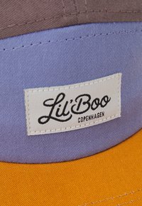 Casquette tricolore avec un tissu violet sur le haut, un visière orange et une étiquette blanche brodée avec "Lil' Boo Copenhagen" en noir.