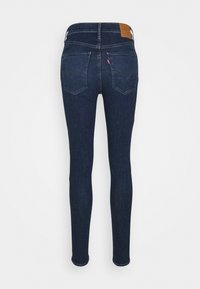 Mörkblå skinny jeans foto tagit bakifrån med två sydda fickor, en liten röd etikett på höger ficka och en brun läderetikett på midjebandet.
